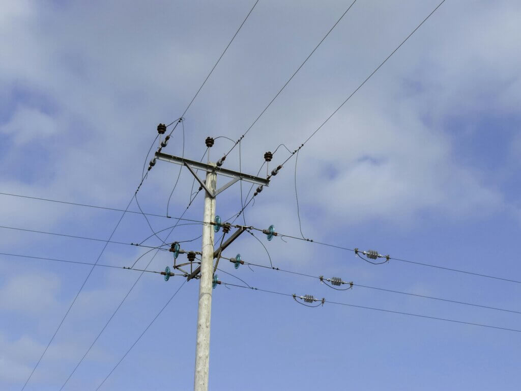 Looking up at a wood utility pole with multiple crossbars, insulators, and power lines spanning across a partly cloudy blue sky.