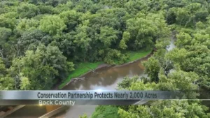 An aerial view of a winding river through dense summer forest in Rock County, with a news chyron reading "Conservation Partnership Protects Nearly 2,000 Acres."