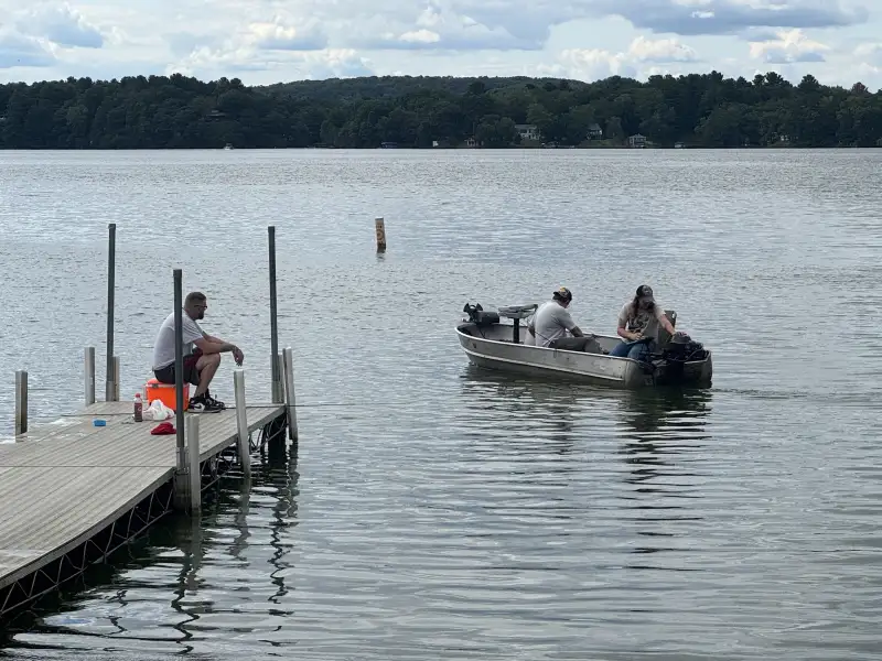 A person sits on a dock watching two others in a small aluminum boat on a calm lake, with a forested shoreline and cottages visible in the distance.